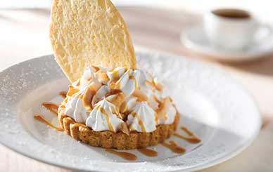 Coconut cream pie topped with swirls of whipped cream and caramel drizzle, served with a crisp coconut tuile on a white plate.