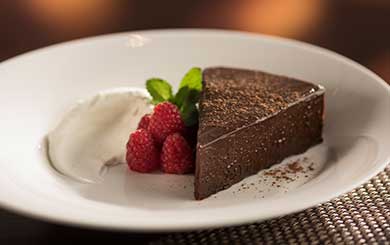 Slice of rich chocolate cake dusted with cocoa powder, served on a white plate with fresh raspberries, a sprig of mint, and a dollop of whipped cream.